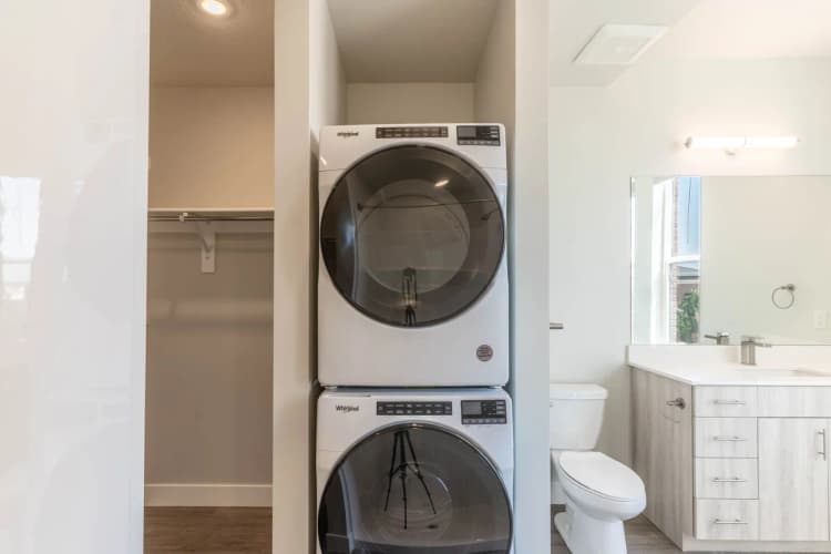 Kenosha apartments for rent - image - Modern laundry area with a sleek stacked washer and dryer beside a bright, elegant bathroom featuring a large mirror and contemporary vanity.